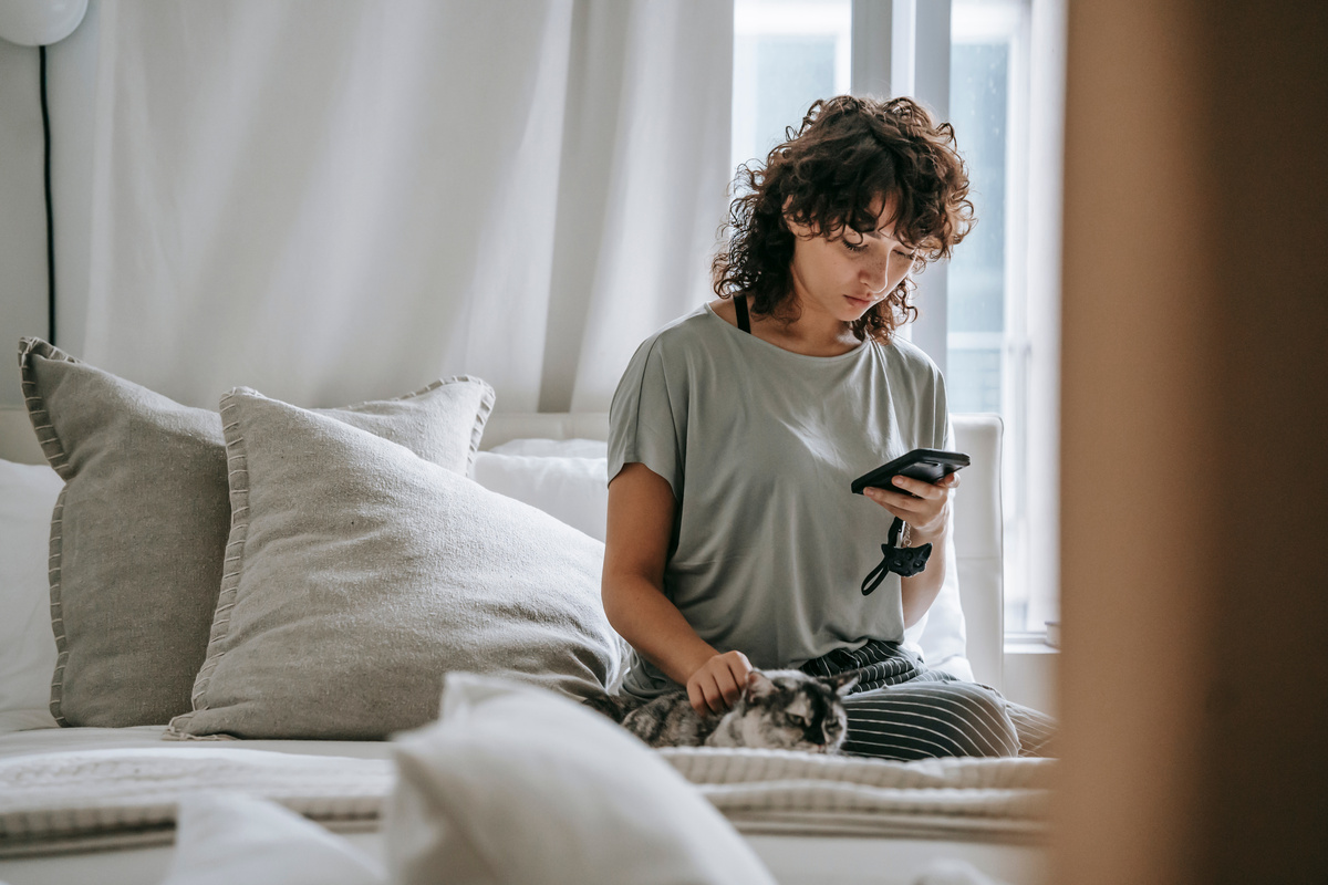 Serious young ethnic lady reading message and petting cat on bed